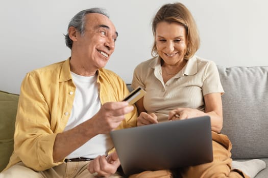 Happy senior spouses shopping online on laptop, man giving credit card to his wife, sitting on sofa at home