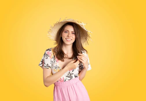Satisfied caucasian smiling lady in summer outfit and straw hat enjoying holiday resort, posing on yellow background