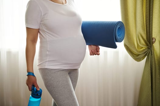 Belly of pregnant woman in late pregnancy, in white mockup t-shirt, holding exercise mat, ready for fitness and yoga
