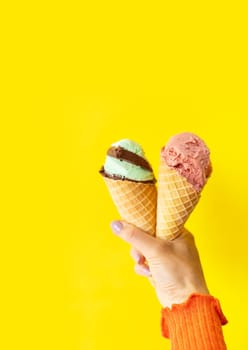 A cone of refreshing ice cream in a waffle cup against a yellow wall. The concept of vacation, vacation at sea. A woman holds ice cream in her hands. Vertical photo.