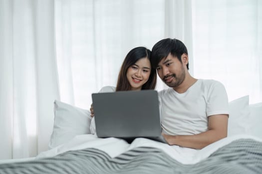 Young couple using laptop sitting on the bed at home. Full length of couple watching movie on laptop. Happy man and woman are relaxing on bed. They are spending leisure time at home