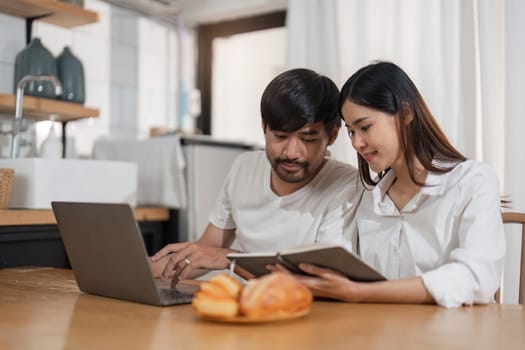 Young asian couple wear casual clothing relaxing while sitting at table in modern kitchen and working with computer laptop at home. Love, happiness, work at home concept