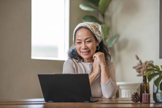 Attractive beautiful mature Asian woman sits at a table with her use laptop in her minimal living room. Hobby and lifestyle concepts..