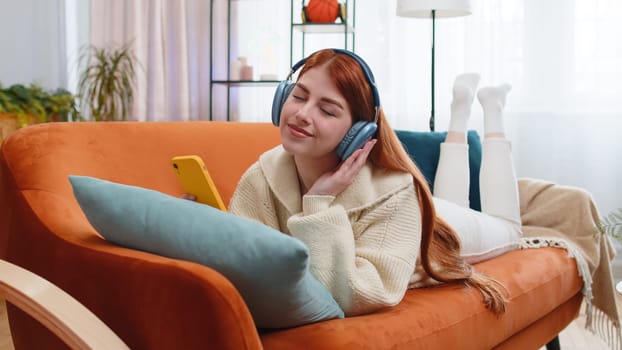 Happy young woman in wireless headphones relaxing lying on sofa at home listening favorite music