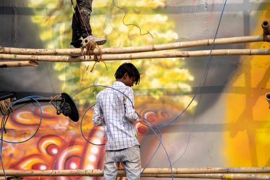 man standing on bamboo scaffolding using a spray air gun to paint a picture of hanuman the hindu god on janaki setu bridge in rishikesh India
