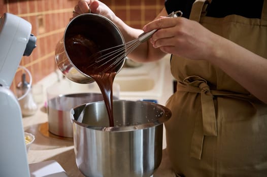 Close-up pastry chef pouring melted chocolate into a planetary mixer bowl, mixing it with cream or whipped egg whites