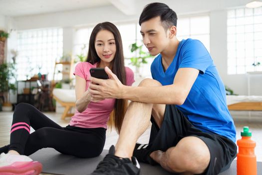 Athletic happy couple sitting on fitness mat, using phone.