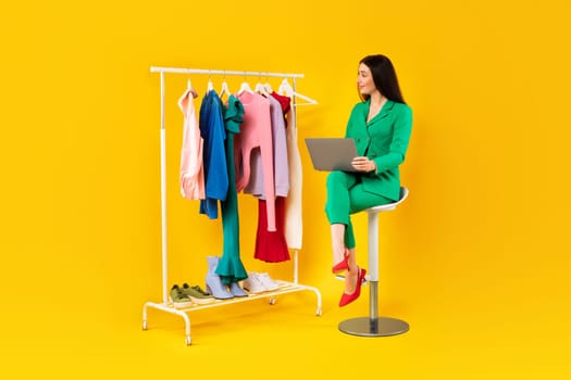 Beautiful woman stylist sitting on yellow background near lot of clothes on garment rack and using laptop, full length
