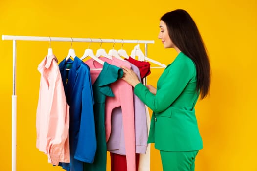 Young stylist woman picking trendy clothing, standing near garment rack, checking new collection, yellow background