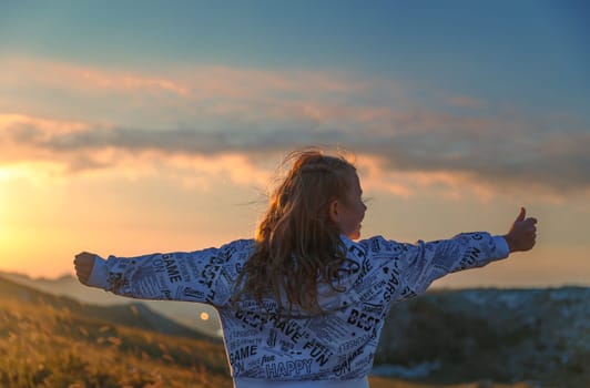 Rear view of a cheerful girl having fun in the mountains. Happy child with raised up hands enjoying mild sunset light. Active summer holidays.