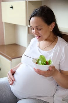 Smiling pregnant woman caressing her belly, eating healthy salad, expressing positive emotions from maternity lifestyle