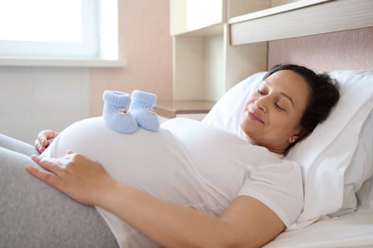 Happy pregnant woman expecting baby, relaxing at home, putting baby booties on her big belly. Pregnancy trimester third