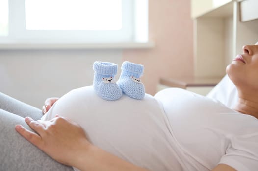Pregnant woman expecting future baby, relaxing at home, putting baby booties on her big belly. Copy advertising space