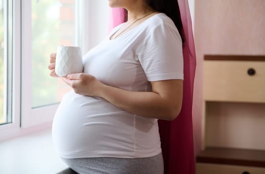 Close-up of a happy pregnant woman standing by window at home bedroom, relaxing with a cup of herbal drink