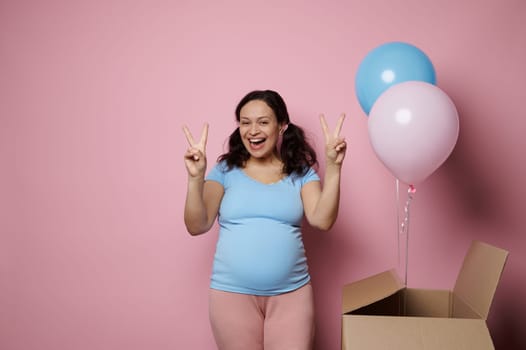 Pregnant woman smiles and shows two fingers at camera, feeling positive emotions expecting twins. Gender reveal party.