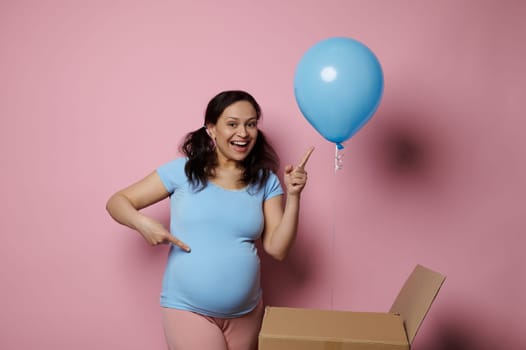 Expectant pregnant woman smiles points at blue balloon, she's expecting a baby boy, pink background. Gender reveal party