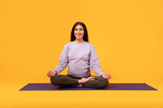 The quiet strength of motherhood. Happy pregnant woman sitting on yoga mat, meditating and smiling, yellow background