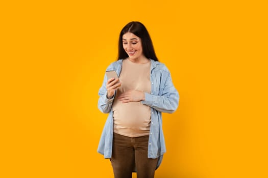 Smiling young pregnant woman using pregnancy tracker app or reading maternity blog online on phone on yellow background