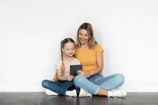 Family Leisure. Happy Young Mother And Cute Little Daughter Using Digital Tablet Together While Sitting On Floor Near White Wall, Smiling Mom And Female Child Shopping Online Or Watching Videos