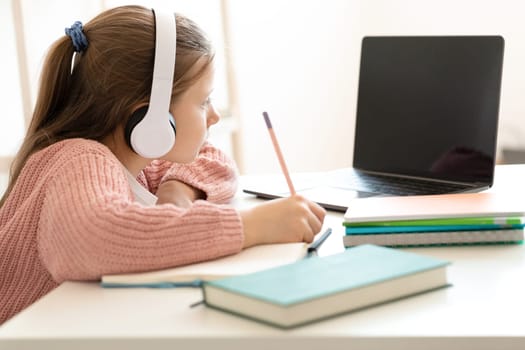 Concentrated caucasian small girl in earphone doing homework, watching video lesson on computer