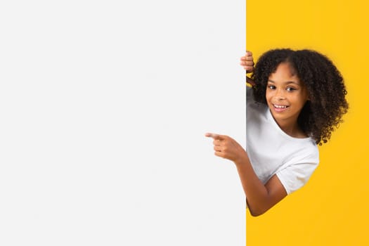 Cheerful adolescent black curly girl in white t-shirt point finger on big banner with empty space