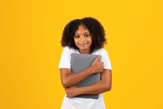 Happy adolescent curly black girl in white t-shirt hugs tablet, isolated on yellow studio background