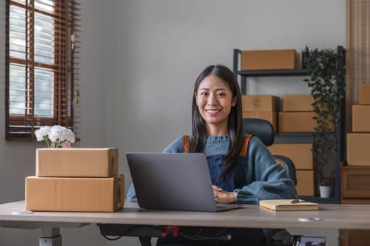 Happy young asian woman startup small business freelance holding parcel box and computer laptop and sitting on chair, Online marketing packing box delivery concept