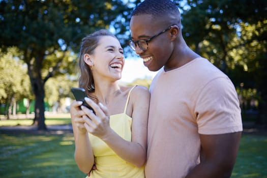 Interracial, social media and couple with a phone in nature, funny communication and laughing at meme. Comic, streaming and black man and woman reading a joke on a mobile in a park in France