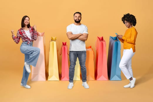 Smiling cheerful beautiful multiracial young people two women and one man enjoying season sale, posing next to big shopping bags on colorful studio background, using cell phone, collage for ecommerce