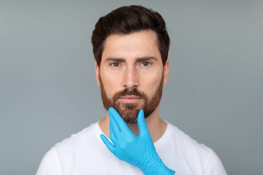 Expert touch. Cosmetician hands in protective medical gloves touching middle aged male face over grey background