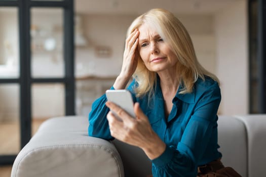 Upset unhappy mature woman using cellphone and touching head, reading bad news, got weird message or email at home
