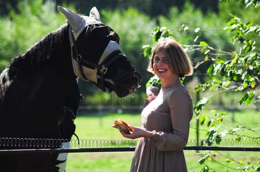 blonde woman feeding a horse among tall trees in summer,countryside, rural