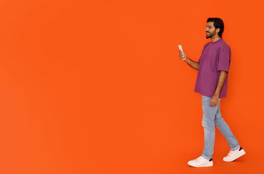 Cool young eastern guy walking with smartphone on orange