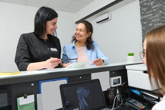 Multi ethnic woman smiles, asking her doctor dentist informations filling in stomatological form, standing by orthodontist reception counter in dentistry clinic. Medical check-up. Healthcare. Medicine