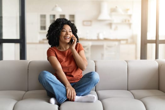 Pleasant Call. Cheerful African American Woman Smiling While Talking On Cellphone At Home, Positive Black Female Enjoying Mobile Phone Conversation While Sitting On Couch In Living Room, Copy Space