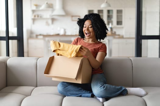 Happy Young Black Woman Unboxing Parcel With New Clothes At Home