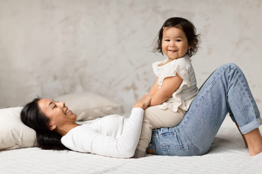 Asian Mother Playing With Baby Daughter Sharing Laughter In Bedroom