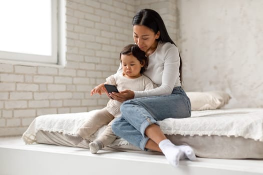 Chinese Mother And Toddler Baby Using Phone In Cozy Bedroom