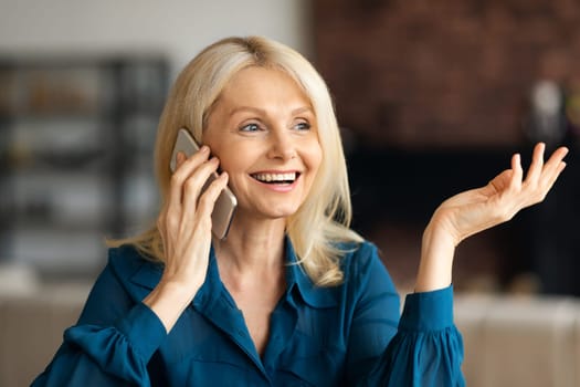 Happy attractive mature businesswoman calling by smartphone, talking and gesturing, sitting in living room interior