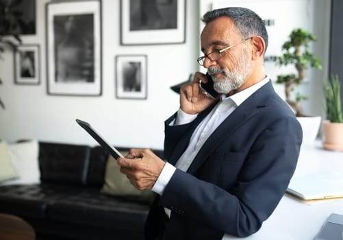 Concentrated european senior businessman in suit, glasses with tablet calls on smartphone in office