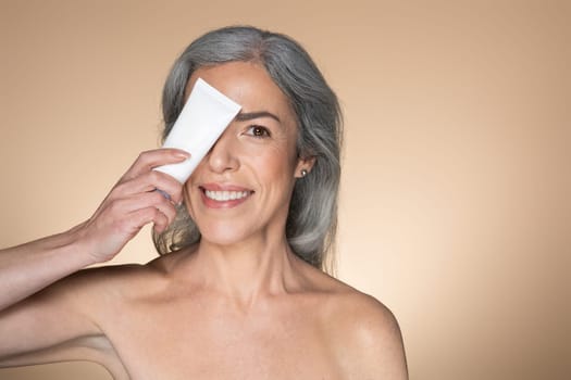Happy grey-haired senior woman holding tube with cosmetic cream near face and smiling, beige background, free space