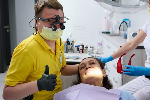 Happy professional male dentist thumbs up while performs examination of a female patient oral cavity in dentistry clinic