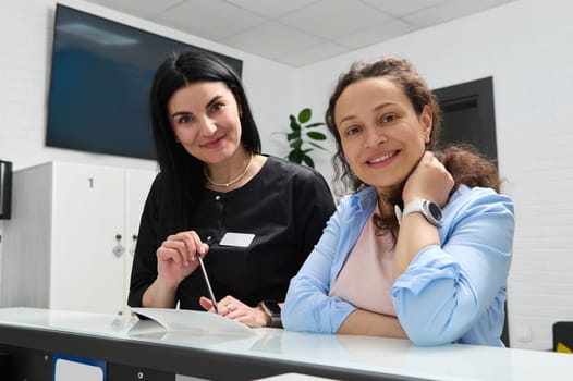 Female dentist and pregnant patient smiling looking at camera, standing at reception counter in modern dental clinic