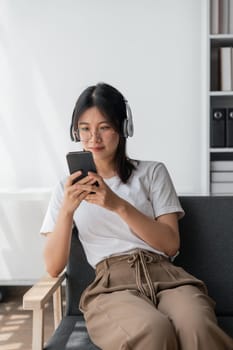 Young Asians woman listening to music on couch in living room at home. Happy Asia female using mobile smartphone, wearing headset and sitting on sofa