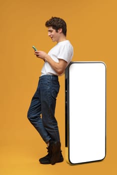 Handsome young man standing leaned on huge smartphone over yellow background