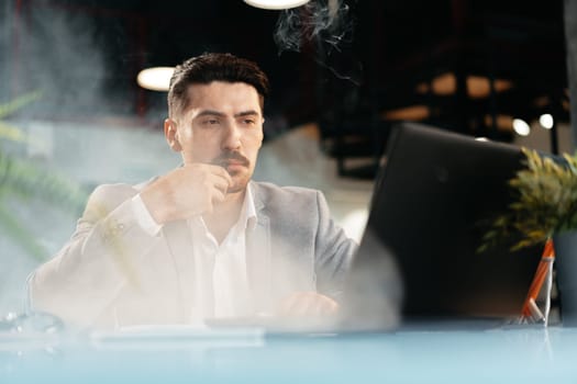 Young businessman smoking an electronic cigarette in his office while working