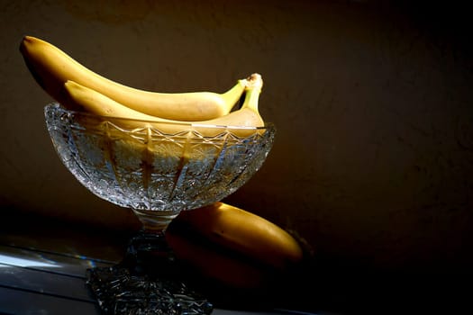 Ripe bananas in a crystal fruit vase salad bowl in sunbeam in dark shadows