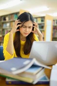 Exhausted female student sitting in library with laptop, suffering from headache or feeling tired of studying for college exam.