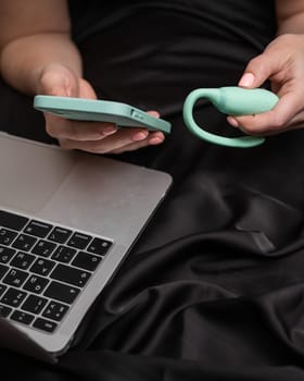 A woman lies on a bed and synchronizes the kegel machine with a smartphone. Girl watching video on laptop.