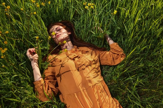 a calm woman with long red hair lies in a green field with yellow flowers, in an orange dress with her eyes closed, spreading her arms to the sides, enjoying peace and recuperating